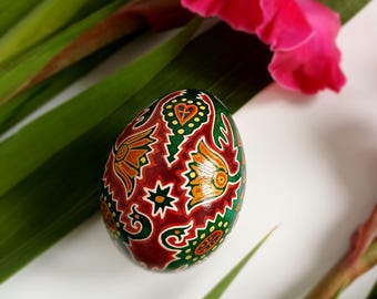 May include: A decorated Easter egg with a maroon background and intricate designs in green, yellow, and orange. The egg is surrounded by green leaves and a vibrant pink flower, creating a festive and colorful composition.