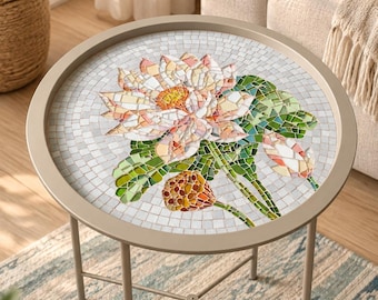 May include: A round side table with a light beige metal frame and a mosaic tabletop. The mosaic features a central lotus flower in shades of pink, white, and yellow, surrounded by green leaves and a brown seed pod, all set against a white background.