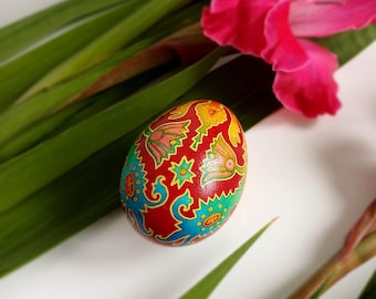 May include: A decorated red Easter egg with floral and geometric patterns in blue, yellow, and green. The egg rests among green leaves and a vibrant pink flower, creating a festive and colorful composition.