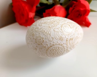 May include: A white, egg-shaped decorative object with intricate, embossed floral and bird patterns. The egg is set against a white surface, with blurred red roses in the background, creating a delicate and artistic composition.