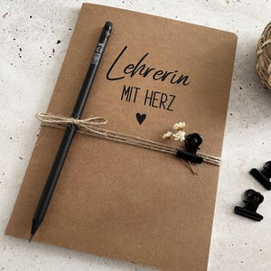 Könnte beinhalten: Ein braunes Notizbuch mit dem deutschen Text "Lehrerin MIT HERZ" und einem kleinen Herzsymbol. Ein schwarzer Bleistift und Bindfaden sind um das Notizbuch gewickelt. Kleine weiße Blumen und schwarze Büroklammern sind am Bindfaden befestigt.