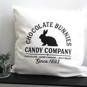May include: White pillow with black text that reads "Chocolate Bunnies Candy Company, Chocolate - Hard Candies - Jelly Beans, Since 1893". A black silhouette of a rabbit is in the center of the text.