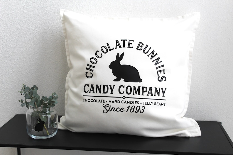 May include: White pillow with black text that reads "Chocolate Bunnies Candy Company, Chocolate - Hard Candies - Jelly Beans, Since 1893". A black silhouette of a rabbit is in the center of the text.