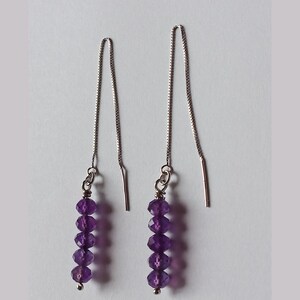 Puede incluir: Un par de pendientes colgantes con una cadena plateada y una serie de cuentas de amatista morada facetadas. Los pendientes tienen un diseño sencillo y elegante con una cadena plateada y un poste que atraviesa la oreja.