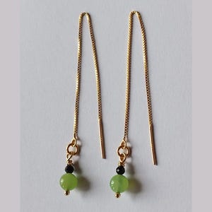 May include: A pair of gold-colored threader earrings. Each earring features a small, light green bead, a black bead, and a gold-colored decorative element. The earrings have a simple, elegant design.