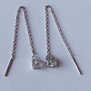 Könnte beinhalten: Ein Paar silberne Ohrhänger mit kleinen, oval geformten, hellgrünen Edelsteinen. Die Ohrringe haben zarte Kettenglieder und einen geraden Stab am Ende jeder Kette. Der Schmuck wird auf einer weißen Oberfläche präsentiert.