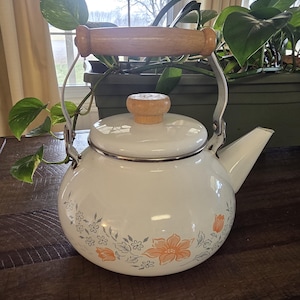 May include: White enamel tea kettle with a wooden handle and lid knob. The kettle features a floral design in orange and gray. The handle is made of wood and the spout is on the right side. The kettle is sitting on a wooden surface.