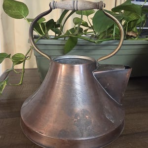 May include: A vintage copper kettle with a wooden handle and brass accents. The kettle has a rounded body and a spout on the side. The handle is made of light-colored wood. The kettle is sitting on a wooden surface.