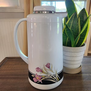 May include: White thermal carafe with a black band and floral design featuring pink and yellow flowers. The carafe has a silver lid and a handle.