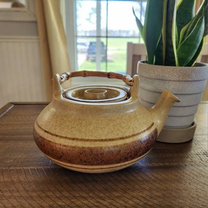 May include: A brown and tan ceramic teapot with a bamboo handle. The teapot has a brown stripe around the middle and a brown rim.
