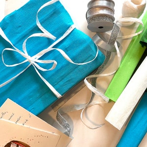 Puede incluir: Un regalo envuelto en papel turquesa atado con cinta blanca. Cintas plateadas y blancas, junto con rollos de papel crep&eacute; verde, blanco y turquesa, est&aacute;n dispuestas cerca. Se puede ver una peque&ntilde;a tarjeta.