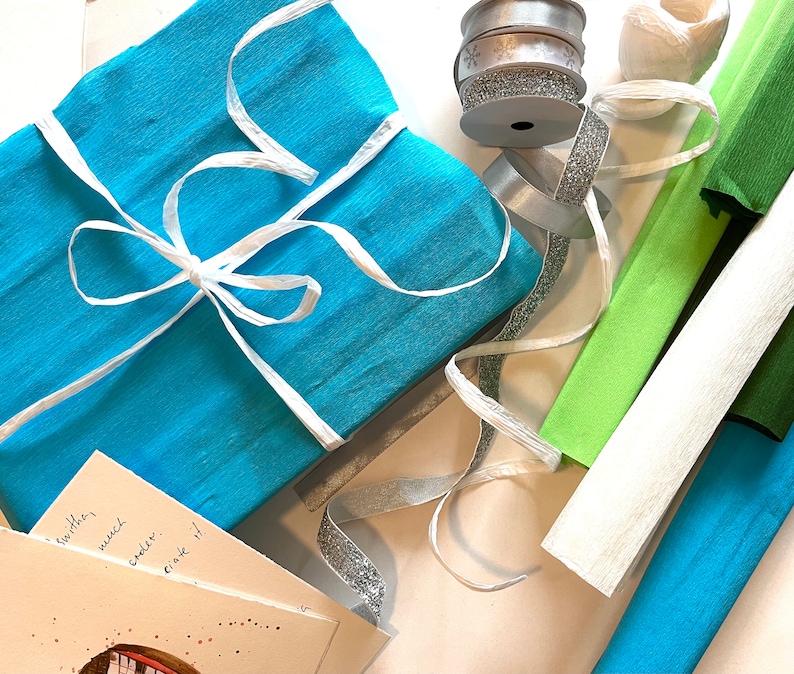 Puede incluir: Un regalo envuelto en papel turquesa atado con cinta blanca. Cintas plateadas y blancas, junto con rollos de papel crep&eacute; verde, blanco y turquesa, est&aacute;n dispuestas cerca. Se puede ver una peque&ntilde;a tarjeta.