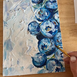 May include: An original painting on canvas featuring a textured white background with blue brushstrokes and a cluster of blue flower blooms. The artist's hand holds a paintbrush, adding the finishing touches to the artwork.