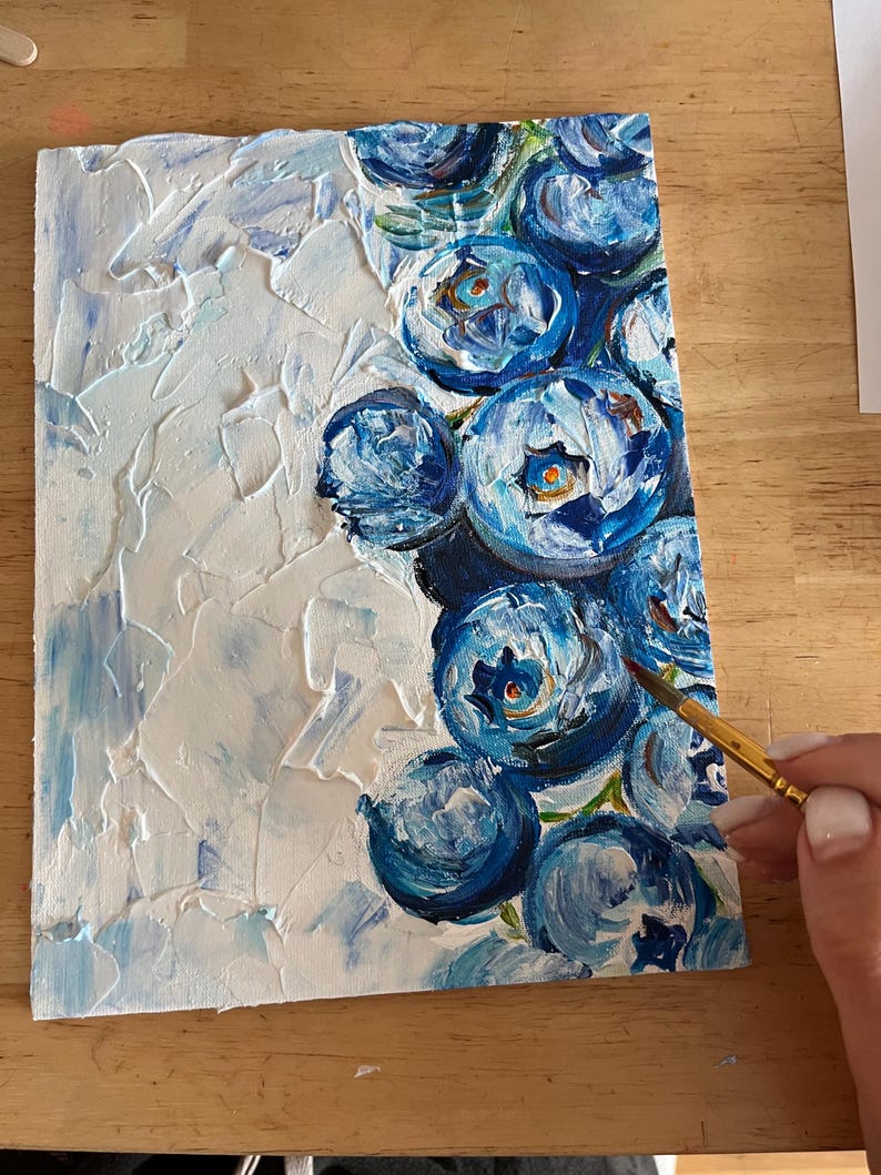 May include: An original painting on canvas featuring a textured white background with blue brushstrokes and a cluster of blue flower blooms. The artist's hand holds a paintbrush, adding the finishing touches to the artwork.