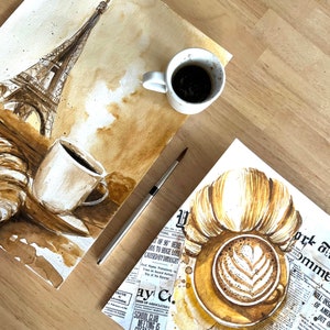 K&ouml;nnte beinhalten: Zwei Aquarellbilder auf Papier, eines zeigt den Eiffelturm und eine Tasse Kaffee, das andere ein Croissant und eine Tasse Cappuccino. Beide Bilder sind in Braun- und Beiget&ouml;nen gehalten, wobei der Kaffee und der Cappuccino realistisch dargestellt sind.