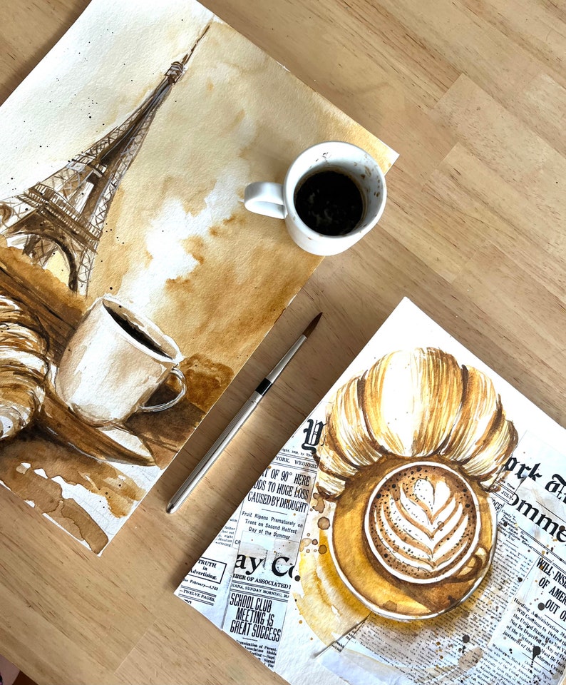 K&ouml;nnte beinhalten: Zwei Aquarellbilder auf Papier, eines zeigt den Eiffelturm und eine Tasse Kaffee, das andere ein Croissant und eine Tasse Cappuccino. Beide Bilder sind in Braun- und Beiget&ouml;nen gehalten, wobei der Kaffee und der Cappuccino realistisch dargestellt sind.