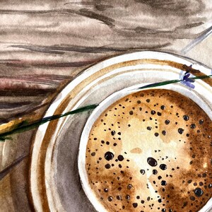 May include: A close-up of a cup of coffee with a lavender sprig on a wooden surface. The coffee is brown and has a foamy top. The lavender is purple and green.
