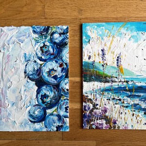 May include: Two small, textured oil paintings. One features a cluster of large, blue blueberries against a white background. The other depicts a coastal landscape with a blue sea, sky, and purple flowers. Birds fly in the sky.