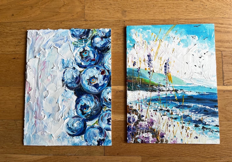 May include: Two small, textured oil paintings. One features a cluster of large, blue blueberries against a white background. The other depicts a coastal landscape with a blue sea, sky, and purple flowers. Birds fly in the sky.