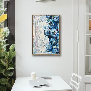 May include: An abstract oil painting of blueberries in a wooden frame. The artwork features a textured white background with a cluster of blue blueberries on the right side. The painting is displayed on a white wall above a white table.