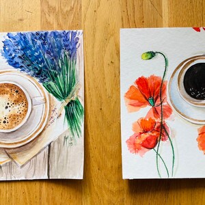 May include: Two watercolor paintings of coffee cups with floral accents. The first painting features a cup of coffee with a lavender bouquet on a wooden surface. The second painting features a cup of coffee with red poppies on a white background.