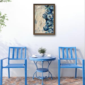 May include: An abstract painting in a brown frame, featuring blue floral shapes against a textured cream background. The painting hangs above a blue patio set with two chairs and a small round table. The table is set with two cups and a small plant.