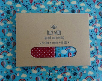 Może przedstawiać: Brązowy papierowy pakiet z tekstem "buzz wrap natural food covering *re-think* *reduce* *re-use*" i logo pszczoły. Pakiet zawiera dwa opakowania do żywności, jedno czerwone w białe kropki i jedno niebieskie z balonem na gorące powietrze i wzorem tęczy.