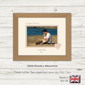 May include: A framed photograph of a father and daughter on the beach. The photo is in a light wood frame with a cream mount. The text "A Daughter's first Love..._ is her Dad" is printed below the photo. The text "Now Double Mounted" is printed below the frame. The text "Orders before 3pm dispatched same day First Class" is printed below the text "Now Double Mounted". The text "Made in the UK" is printed below the text "Orders before 3pm dispatched same day First Class". A small Union Jack flag is printed to the right of the text "Made in the UK".