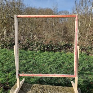 May include: A wooden frame with a red and white striped tape border. The tape reads "PET GRIPPER", "REGULAR CARPET GRIPPER", and "REGULAR CARPET GRIPPER". The frame is standing on a stone surface with green grass in the background.