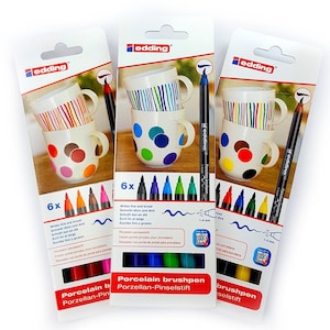 May include: Set of 18 edding porcelain brush pens in various colours. The pens are packaged in three boxes, each with a mug decorated with colourful dots and stripes. The boxes feature the text "Porcelain brushpen" and "Porzellan-Pinselstift".
