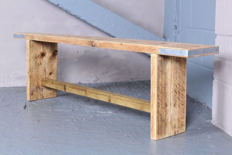 May include: A rustic wooden bench with a natural wood finish and metal accents. The bench has a simple design with a rectangular seat and two legs on each side. The bench is made from reclaimed wood and has a distressed look.