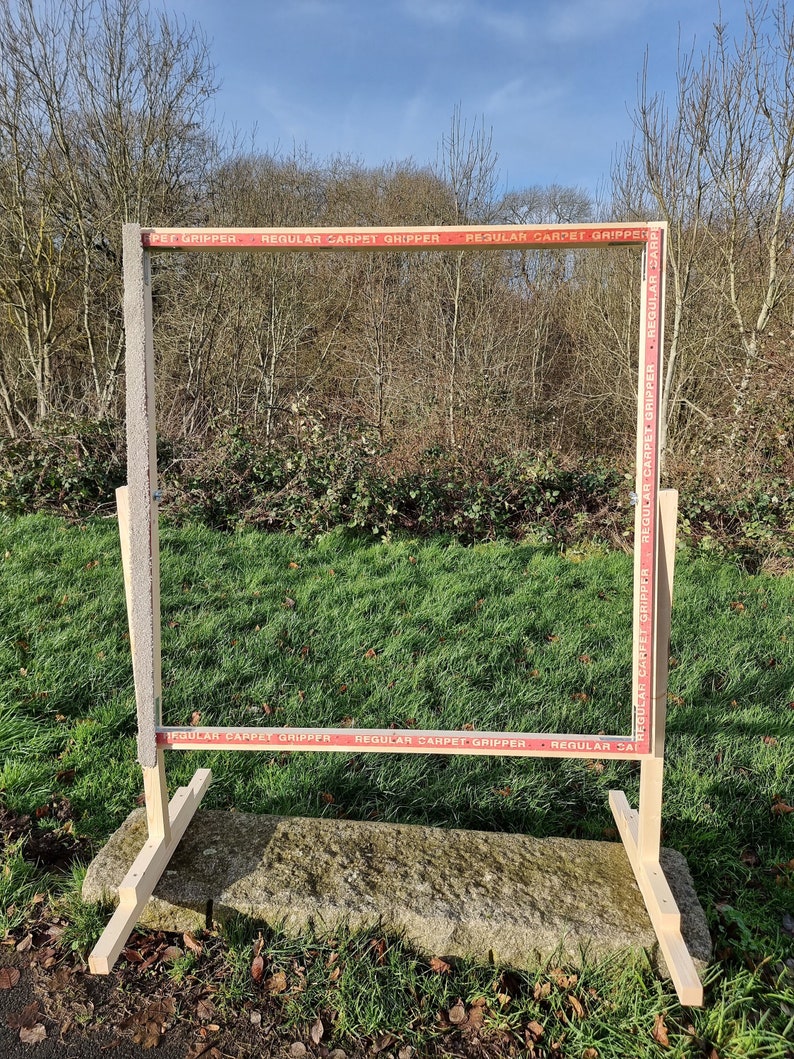 May include: A wooden frame with a red and white striped tape border. The tape reads "PET GRIPPER", "REGULAR CARPET GRIPPER", and "REGULAR CARPET GRIPPER". The frame is standing on a stone surface with green grass in the background.