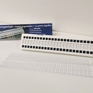 May include: White plastic needle organiser with 50 compartments for storing needles. The organiser comes with 10 blank code cards. The box reads "Needle organiser. Nadel-organisator. Le porte-aiguilles" and "art. nr. 700.050".