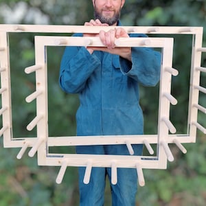 May include: Two wooden frames with dowels around the perimeter. The frames are nested inside each other. A person is holding the frames in front of a blurred green background.