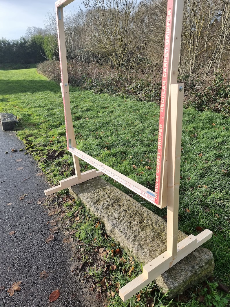 May include: A wooden frame with a red and white label that reads "Regular Carpet Gripper" and "Regular Carpet Gripper". The frame is standing on a stone surface with grass in the background.