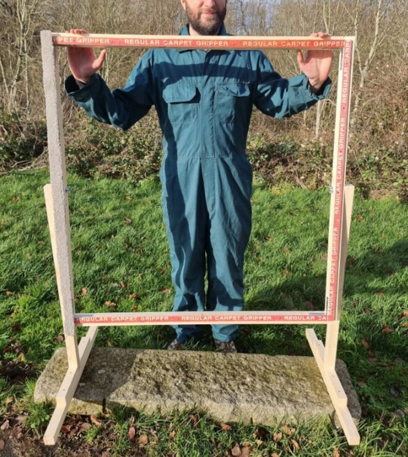 May include: A wooden frame with a red and white striped tape that reads "REGULAR CARPET GRIPPER" is standing on a stone surface. The frame is supported by two wooden legs.