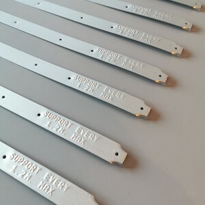 May include: A close-up of eight silver metal strips with the text "SUPPORT EVERY 1.2M. HAX" printed on each strip. The strips are arranged in a staggered pattern.