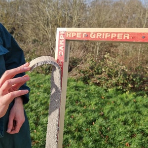 May include: A close-up of a wooden frame with a gray carpet gripper attached to it. The frame is labeled "Carpet Gripper" in red letters.