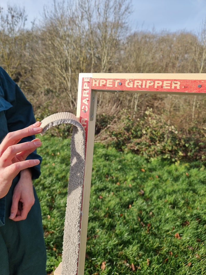 May include: A close-up of a wooden frame with a gray carpet gripper attached to it. The frame is labeled "Carpet Gripper" in red letters.