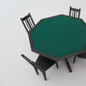 May include: Miniature black table with a green felt top, surrounded by three matching chairs. The table has an octagonal shape and is supported by a single central leg. The chairs have a classic design with vertical back slats.