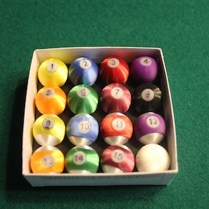 Puede incluir: Un juego de 15 bolas de billar en una caja blanca. Las bolas están numeradas del 1 al 15 y son de varios colores, incluyendo rojo, amarillo, azul, verde, morado y negro.