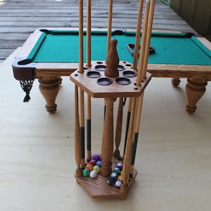 Puede incluir: Un estante de taco de billar de madera con una base hexagonal y un poste central. El estante tiene capacidad para ocho tacos y tiene un compartimento para bolas de billar. El estante está lleno de bolas de billar de colores.