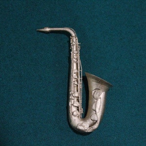 Pode incluir: Um saxofone prateado com um design decorativo na campana. O saxofone está sobre um fundo azul esverdeado.