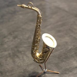 Pode incluir: Um saxofone em miniatura dourado exposto em um pequeno suporte de três pernas. O saxofone tem chaves detalhadas e uma campânula, e é um item decorativo. O suporte é feito de metal fino cor de cobre.