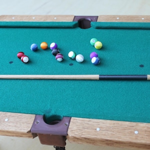 May include: A green felt pool table with a wooden frame and a cue stick. There are 13 multi-colored billiard balls on the table.