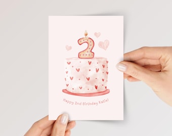 May include: A rectangular greeting card with a light pink background featuring a watercolor illustration of a cake topped with a number two candle. The cake is decorated with small red hearts and dots. The text reads Happy 2nd Birthday Katie.