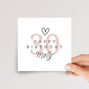 May include: A square white greeting card featuring a large, pale pink number 30 in the background. Centered text reads HAPPY BIRTHDAY in black capital letters, with the name Molly written below in a black cursive script and a small heart icon above.