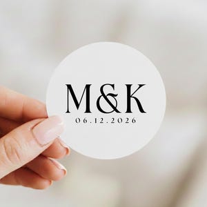 May include: A white, circular sticker with the initials "M&K" in a classic serif font, and the date "06.12.2026" below. The sticker is held by a hand with manicured nails, suggesting a wedding or event theme.