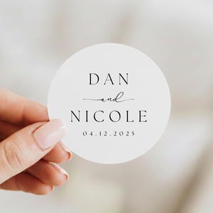 Può includere: Un adesivo rotondo bianco con i nomi "DAN and NICOLE" in nero, con un carattere elegante. Sotto i nomi c'è la data "04.12.2025". L'adesivo è tenuto in mano.