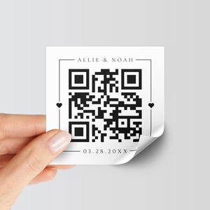 May include: A square white sticker featuring a black QR code in the center. The text ALLIE & NOAH is printed at the top, and the date 03.28.20XX is printed at the bottom. Small black hearts are positioned on the left and right sides of the QR code.